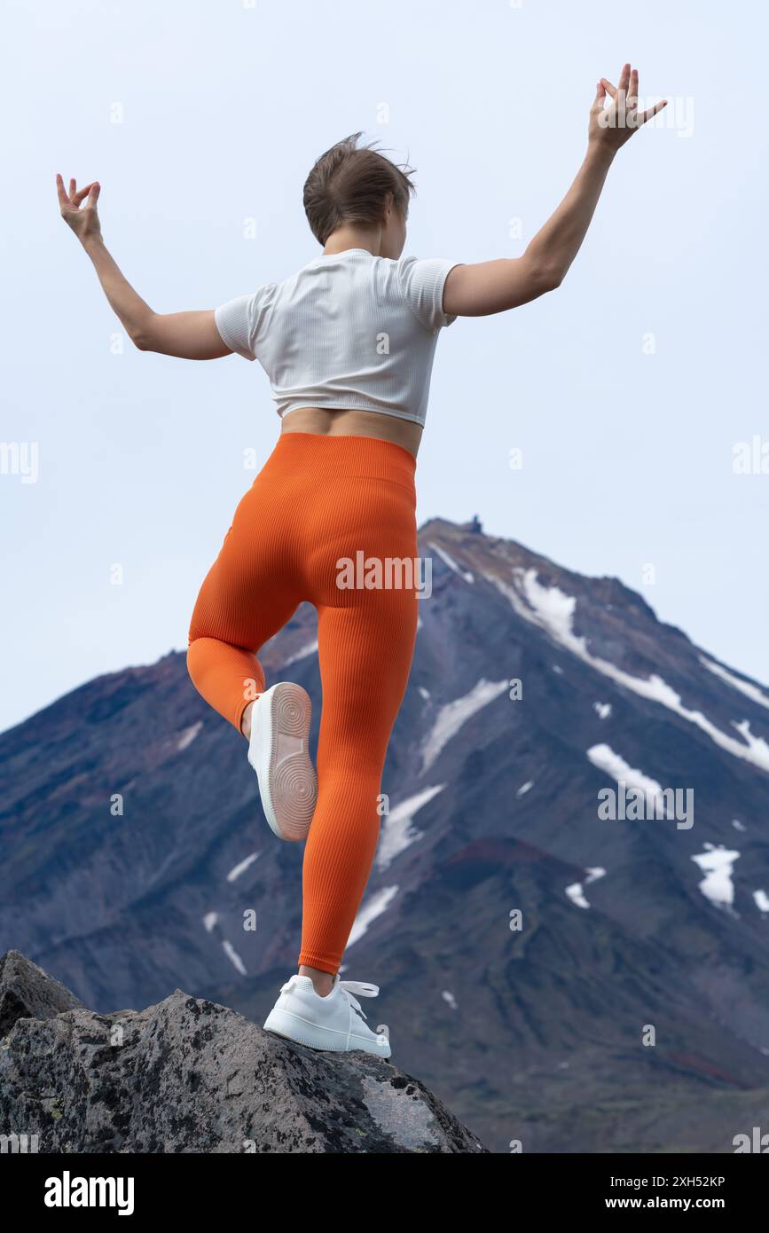 Vista posteriore mozzafiato di una istruttrice di yoga in piedi sulla cima di una montagna rocciosa, bilanciando il suo corpo su una gamba mentre fa yoga con il vulcano Foto Stock