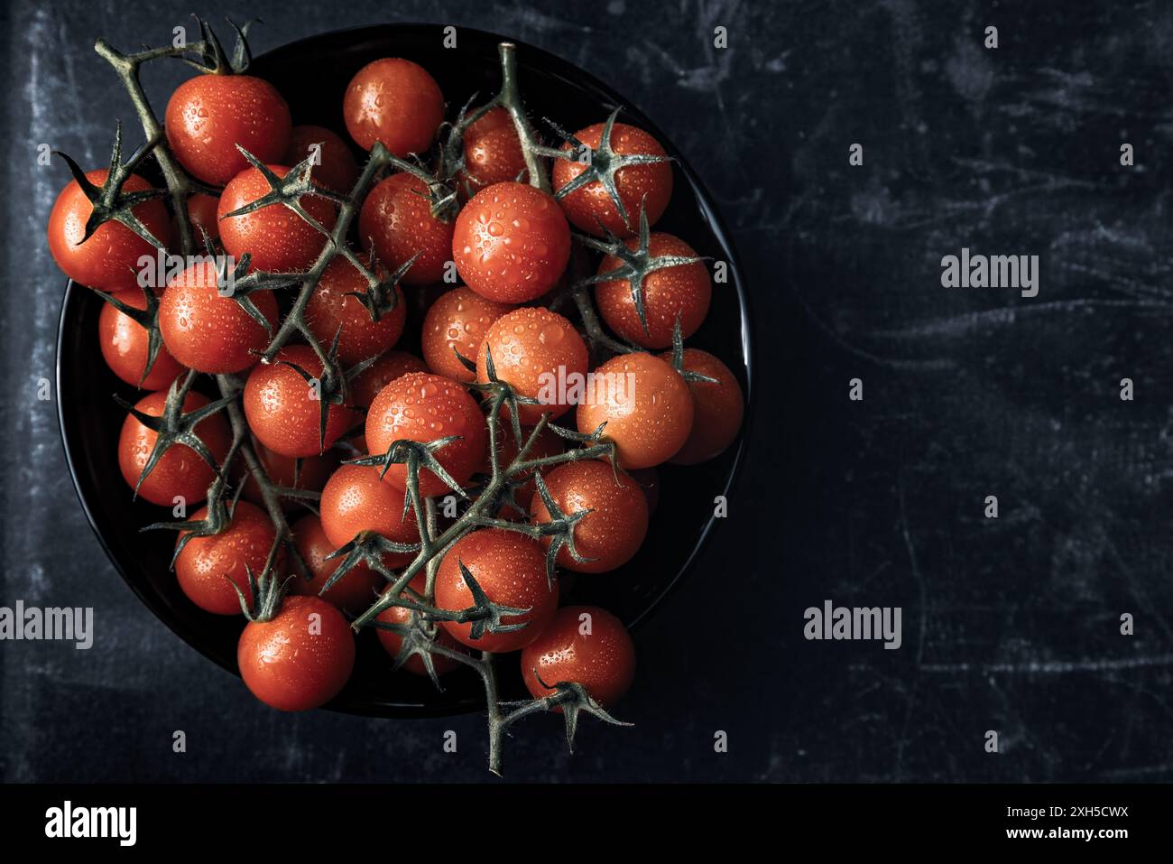Una ciotola nera è piena di pomodori ciliegini freschi e vivaci attaccati alle viti verdi. Lo sfondo scuro e strutturato contrasta con i frutti luminosi. Foto Stock
