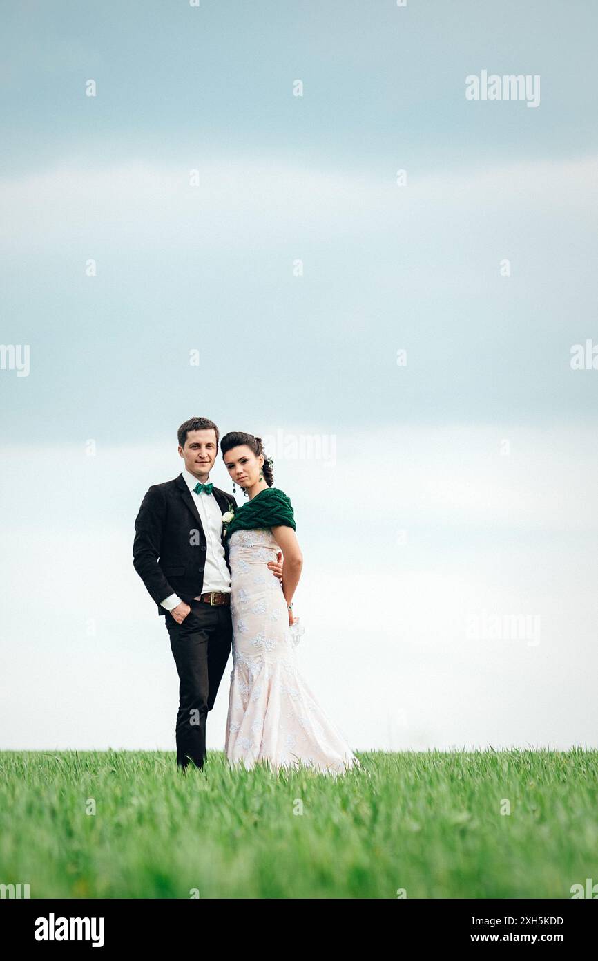 lo sposo in tuta marrone e la sposa in abito color avorio su un campo verde che si stende in lontananza contro il cielo Foto Stock