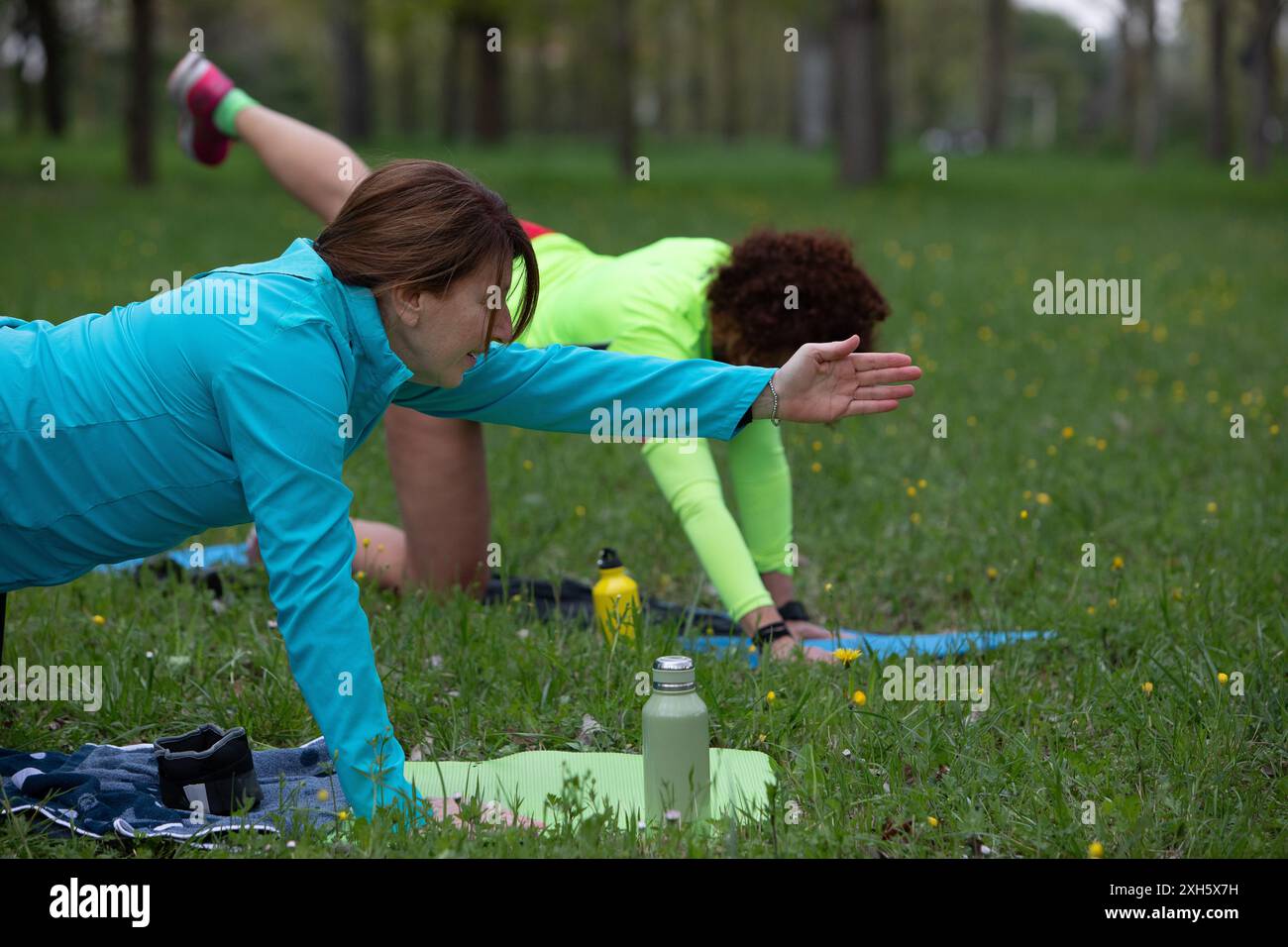 Due donne che indossano abbigliamento sportivo praticano yoga in un parco pubblico sui loro colorati tappetini per yoga, godendosi l'aria fresca e l'erba verde Foto Stock