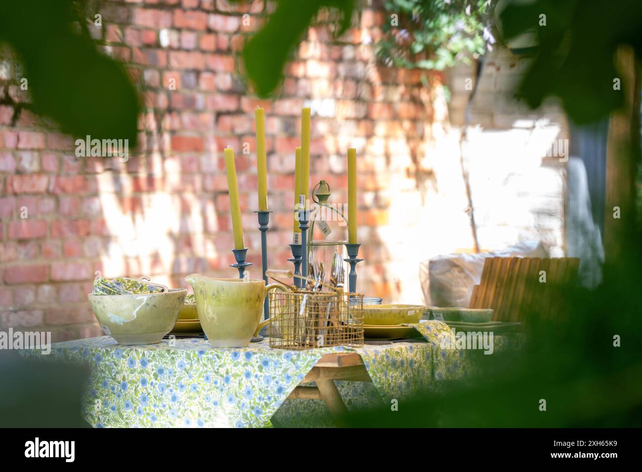 Piatti, ciotole e posate fanno nascere alte candele verdi in candelabri blu su una tovaglia verde e blu girata all'esterno attraverso le foglie verdi vicine Foto Stock