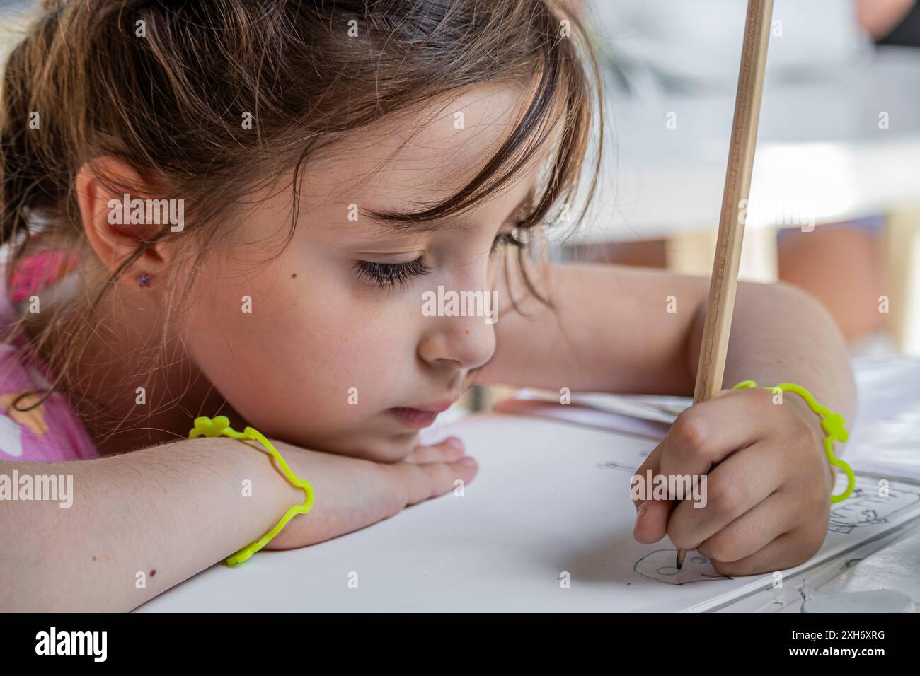 Una piccola e graziosa bambina, disgustata e sudata in una calda giornata estiva, è impegnata a disegnare con matita e pastelli su un foglio bianco. Era seduta ad una Foto Stock