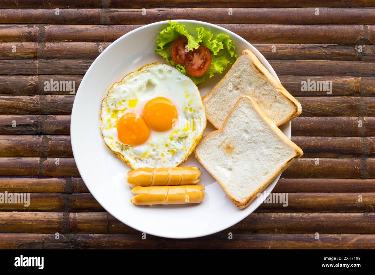 Uova strapazzate, salsicce, pomodoro fresco con insalata verde e toast su piatto bianco su tavola di bambù marrone. Colazione all'americana. Vista dall'alto. Foto Stock