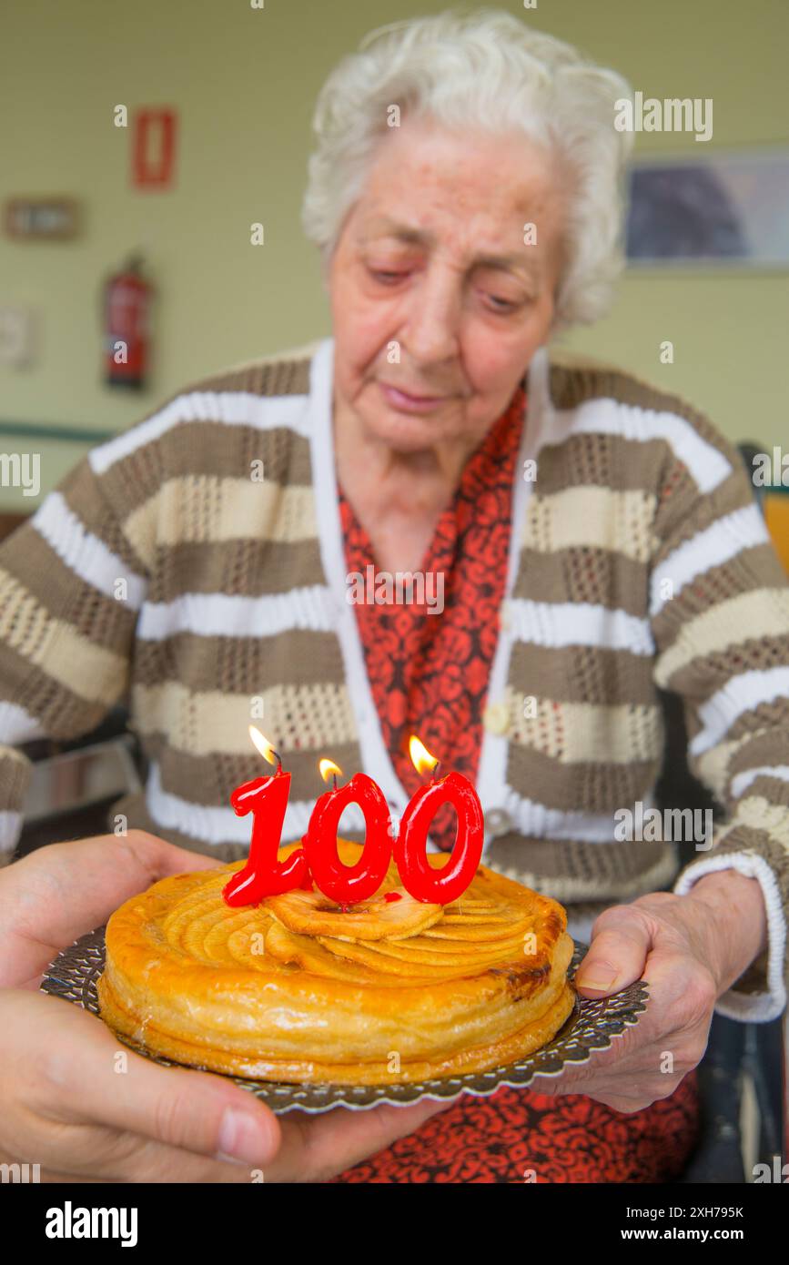 Vecchia donna in una casa di cura, su di lei un centinaio di compleanno, soffiando compleanni candele su una torta detenute da un uomo con le mani in mano. Foto Stock