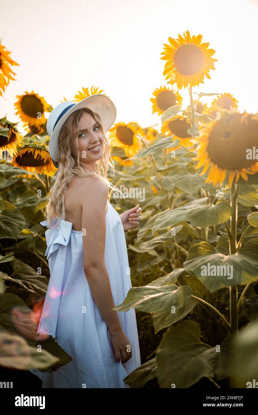 Una donna con un vestito bianco sorride alla macchina fotografica mentre è in piedi in un campo di girasoli. Indossa un cappello di paglia e il sole sta tramontando nel dorso Foto Stock