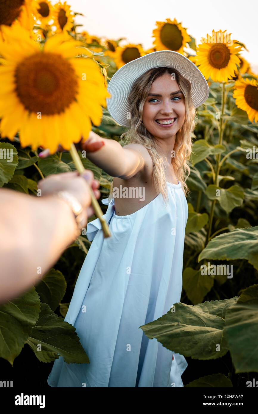 Una donna con un vestito bianco e un cappello di paglia sorride mentre tiene un girasole in un campo di girasoli in fiore. Foto Stock
