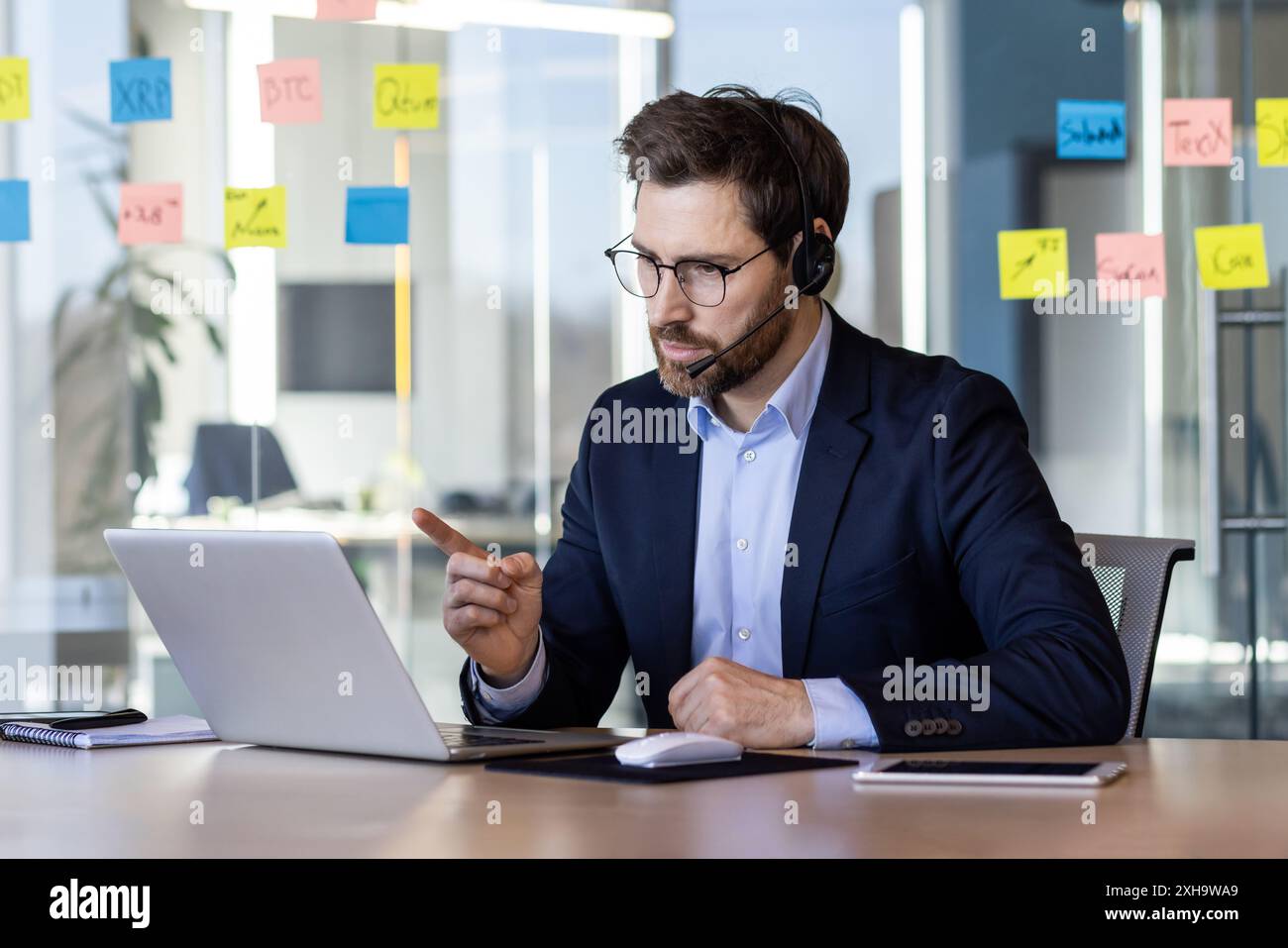 Uomo serio e sicuro di sé con cuffia, telefono e laptop che utilizza videochiamate per le comunicazioni remote, investitore in riunione remota che spiega il successo Foto Stock