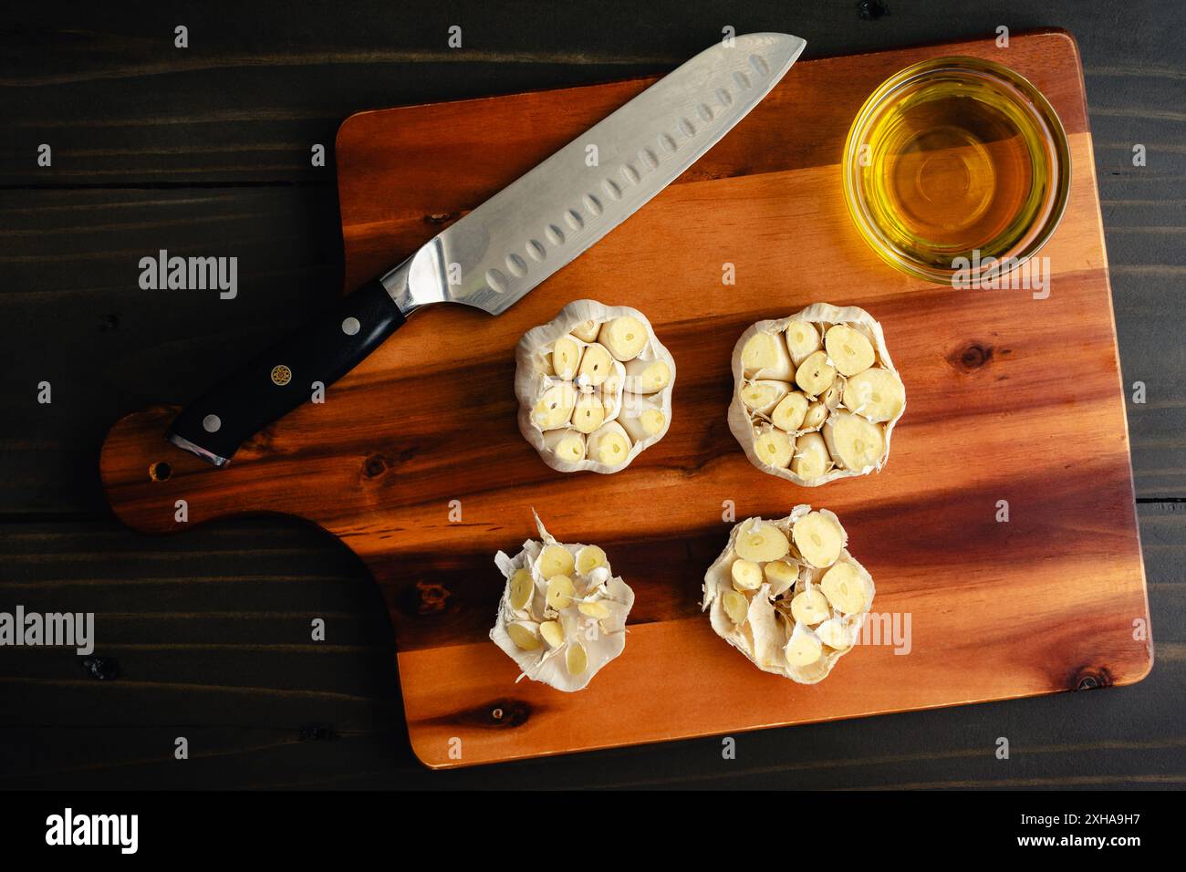 Preparazione dei bulbi d'aglio da tostare in olio d'oliva: Teste d'aglio fresche con il fondo tagliato su un tagliere di legno Foto Stock
