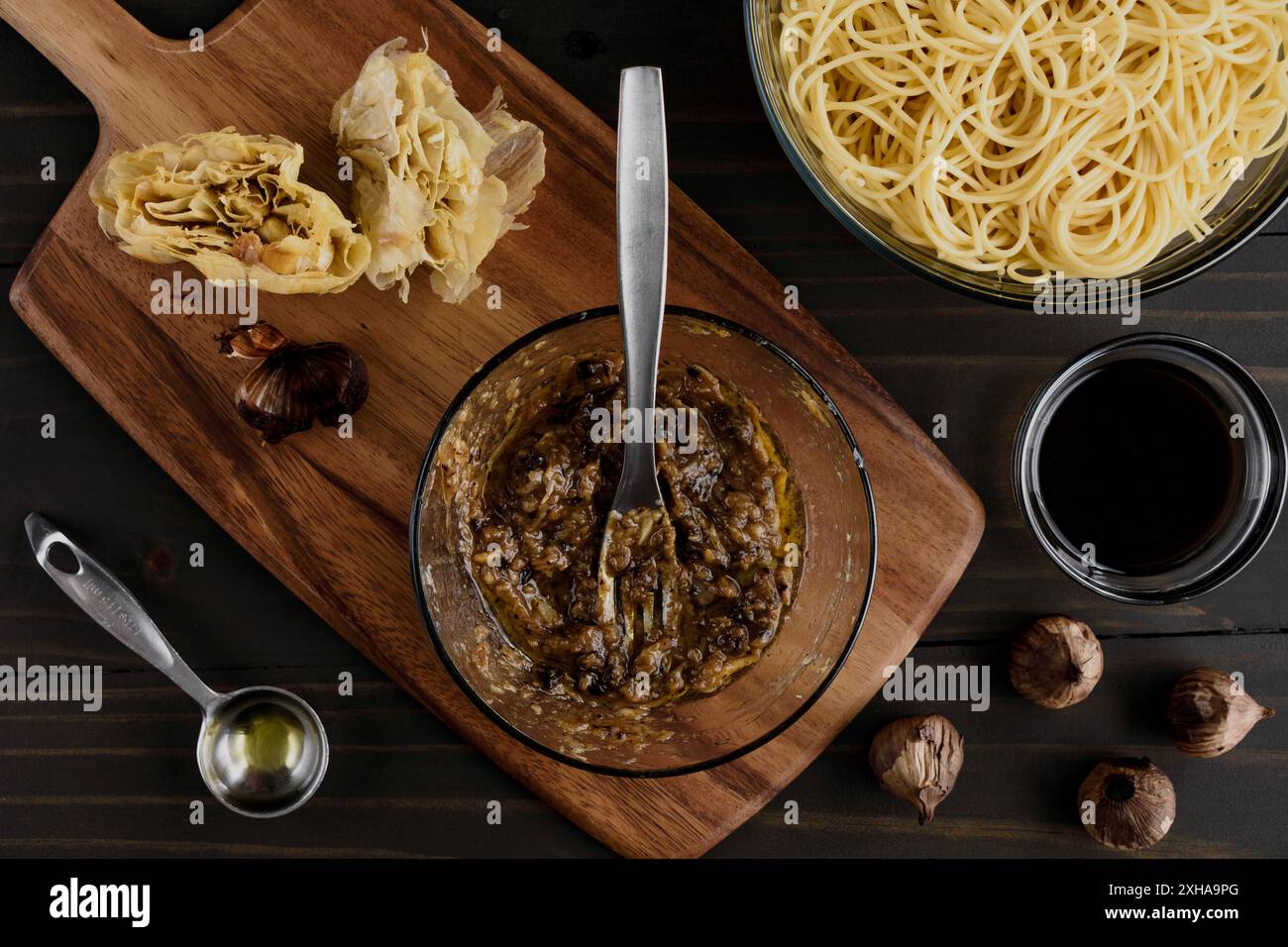 Aglio arrosto e aglio nero schiacciato insieme a una forchetta: Purea di aglio con una ciotola di spaghettini e un piccolo piatto di salsa sullo sfondo Foto Stock