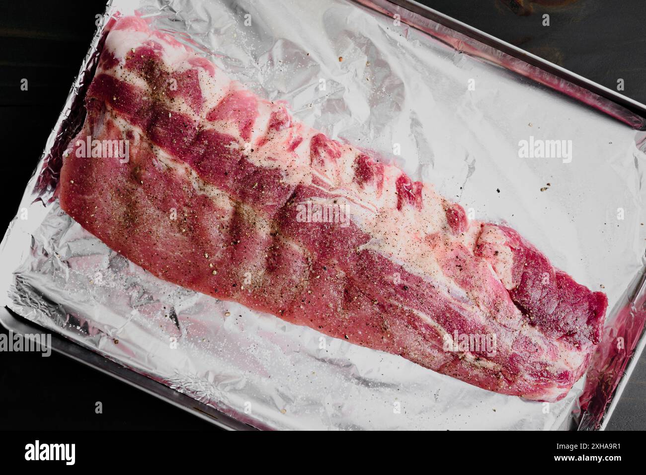 Lastra cruda di costolette di maiale condite con sale e pepe: Griglia di costolette di maiale non cotte su una teglia coperta di foglio di alluminio Foto Stock