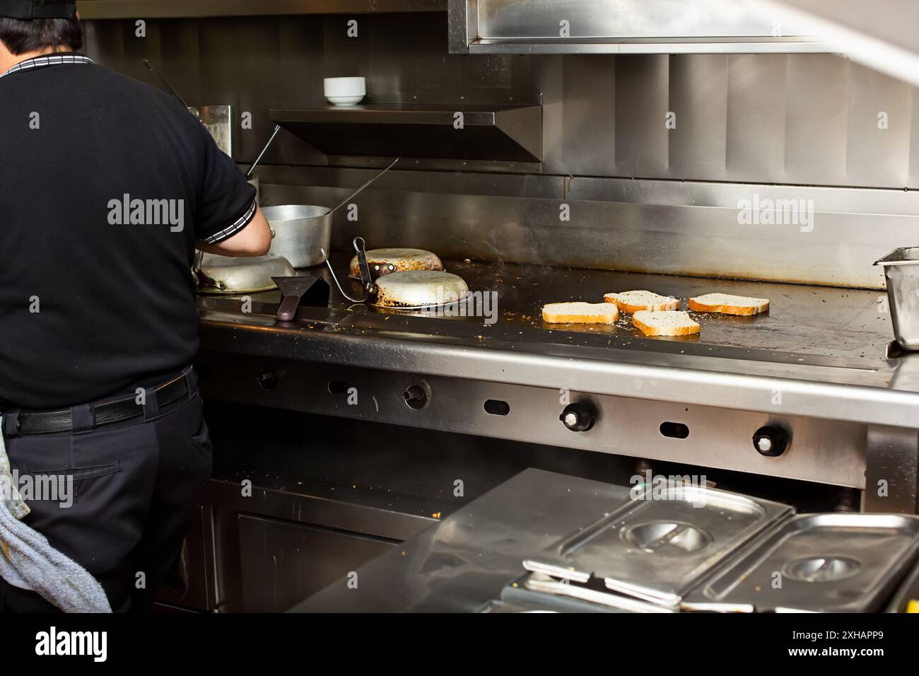 Vista di un cuoco che frequenta un piano cottura e un'area per grigliate. Le fette di pane vengono tostate sulla piastra, in un ambiente di cucina del ristorante. Foto Stock