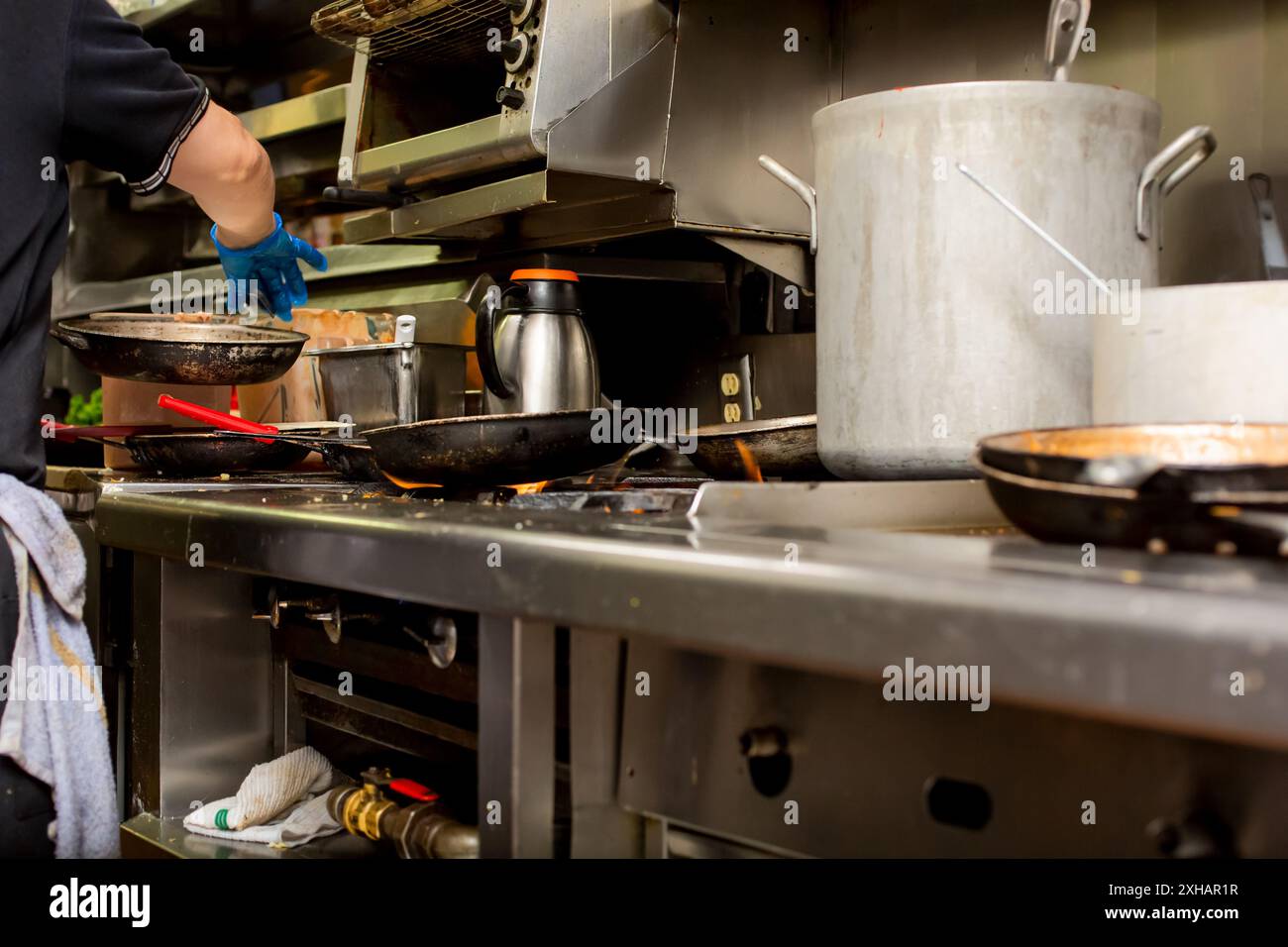 Una vista dall'angolo basso dell'area cucina del ristorante, con pentole, piatti alla griglia, disordine e un cuoco affollato. Foto Stock