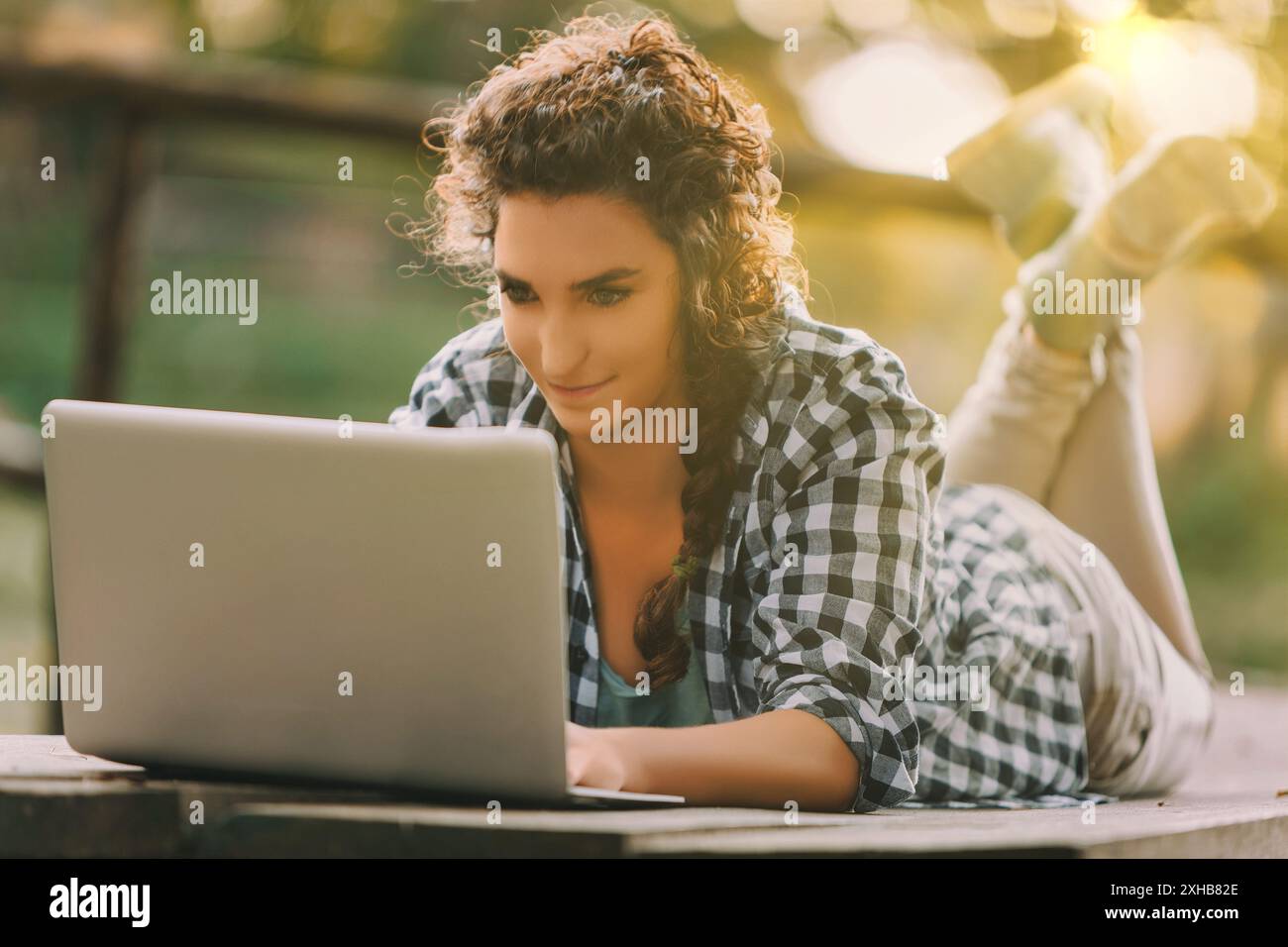 Donna dai capelli ricci con treccia e camicia a scacchi giace sullo stomaco utilizzando un laptop. La sua espressione focalizzata riflette un profondo coinvolgimento, completato Foto Stock