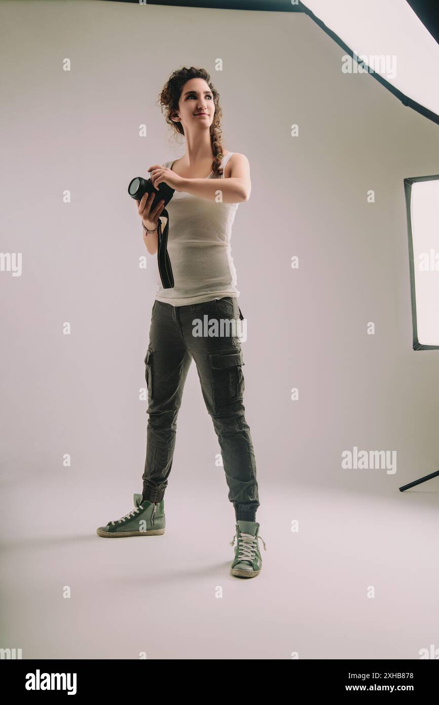 Una giovane donna con i capelli ricci intrecciati, vestita con una capote senza maniche e pantaloni cargo, tiene una macchina fotografica in uno studio. La sua posizione sicura e il suo rifiuto di espressione Foto Stock