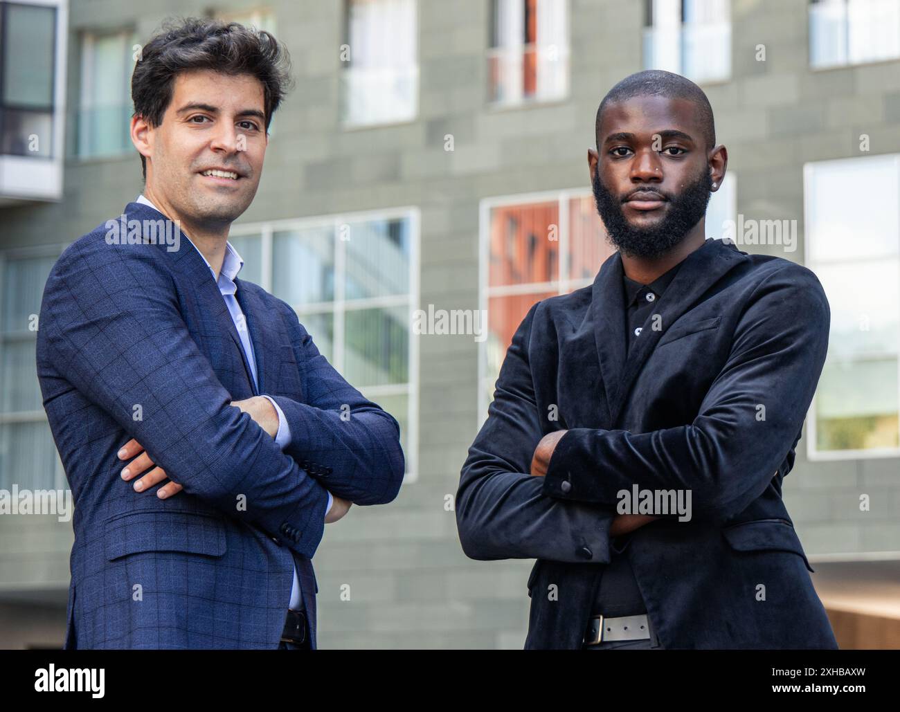 primo piano uomo d'affari nero africano e una controparte ispanica, entrambi vestiti impeccabilmente in abiti, stanno con le braccia incrociate, trasudando fiducia e promesse Foto Stock