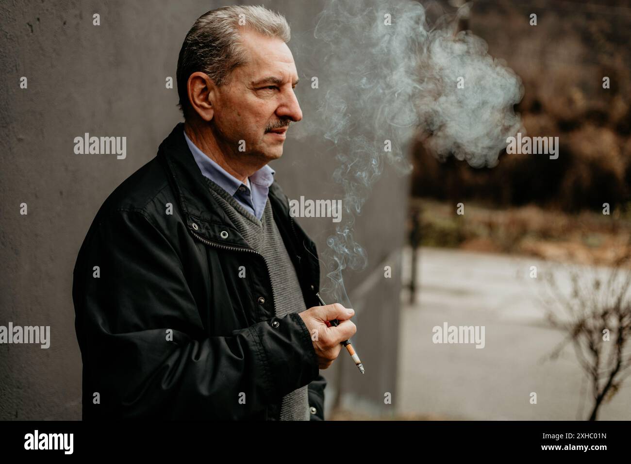 Un uomo anziano con una giacca nera e una camicia blu, sta fuori al freddo, espirando fumo da una sigaretta. Foto Stock