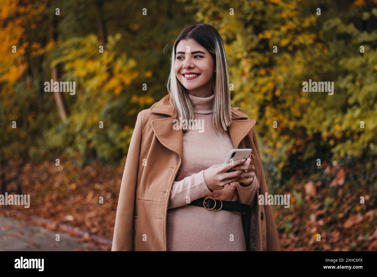 Una donna con un cappotto marrone si trova in un parco autunnale, sorridendo e guardando lateralmente tenendo in mano uno smartphone. Foto Stock
