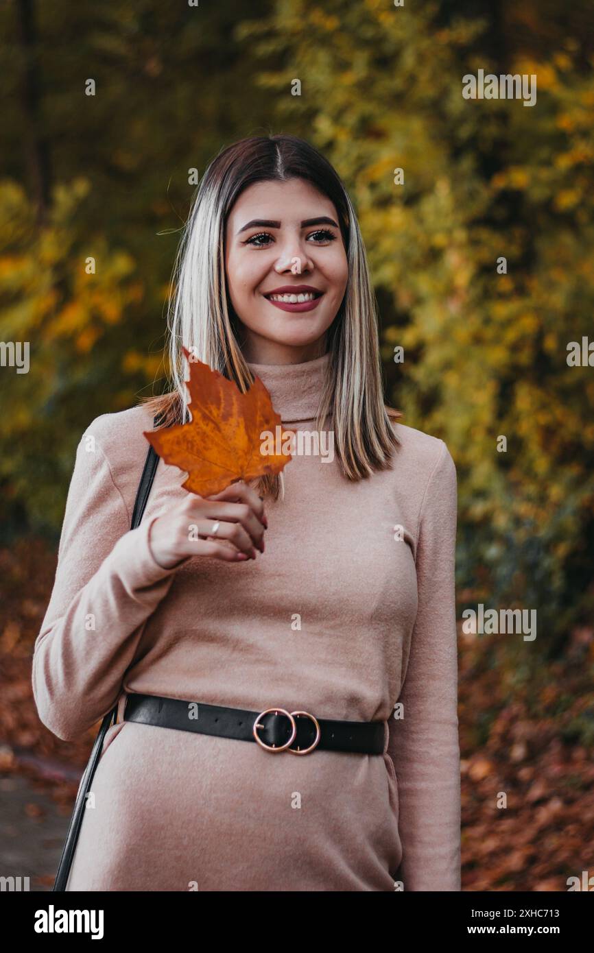 Una donna sorride mentre tiene una grande foglia d'arancia in un parco. Indossa un abito rosa chiaro a maniche lunghe con cintura nera. Foto Stock