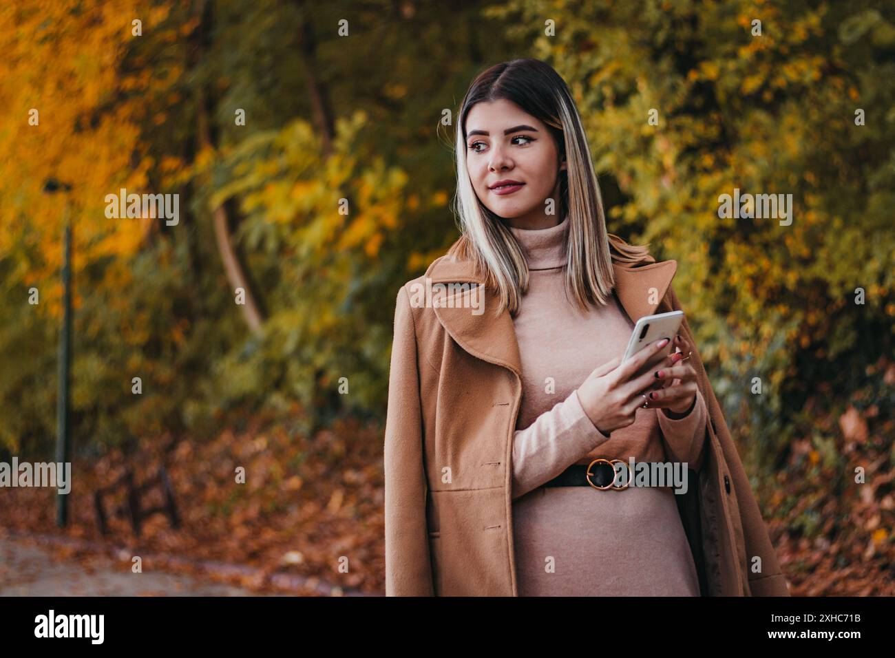 Una donna con un cappotto marrone si trova in un parco con foglie autunnali per terra. Sta tenendo uno smartphone tra le mani e guardando di lato. Foto Stock