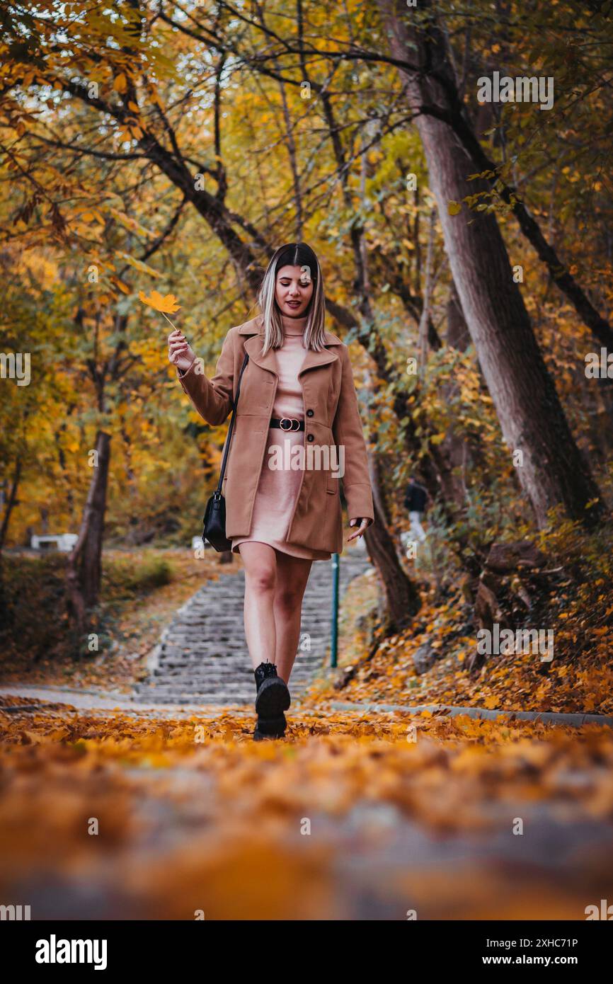 Una donna con un cappotto marrone cammina lungo un sentiero coperto di foglie cadute, circondato da colorati alberi autunnali. Foto Stock