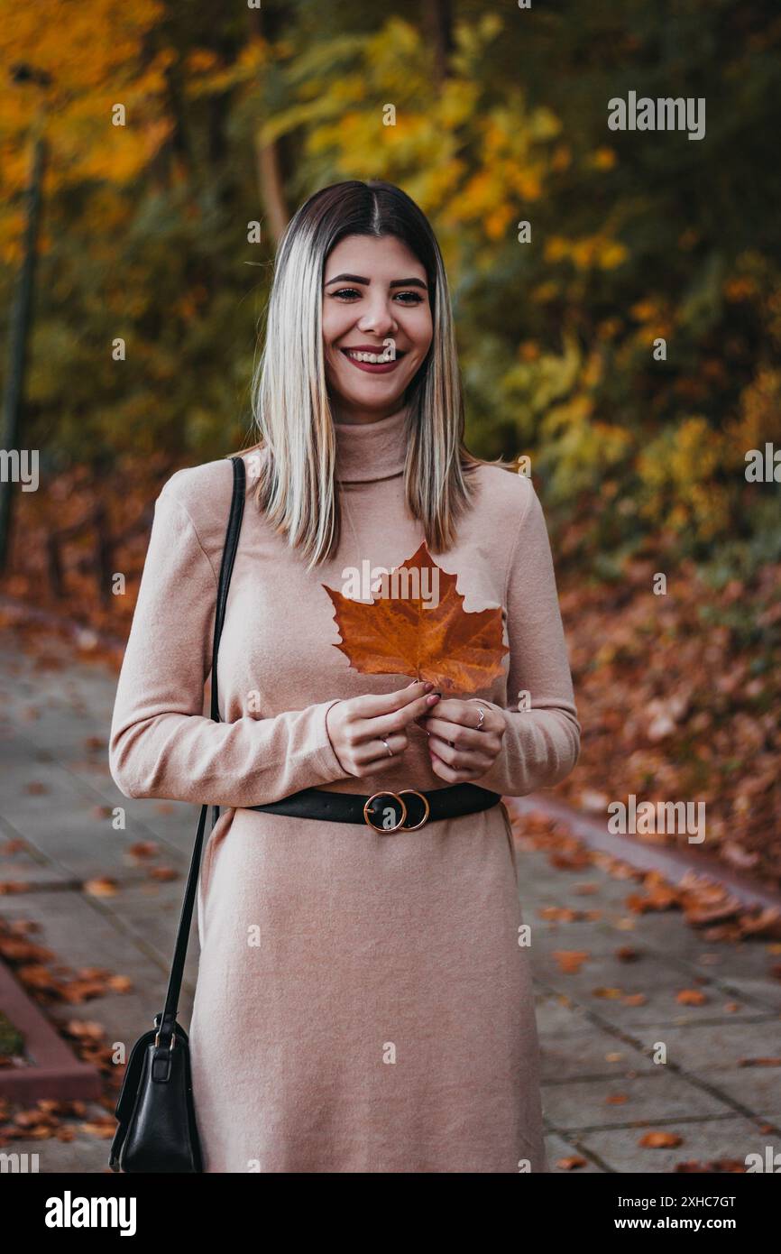 Una donna con un abito rosa a maniche lunghe sorride mentre tiene una foglia marrone in un parco. Foto Stock