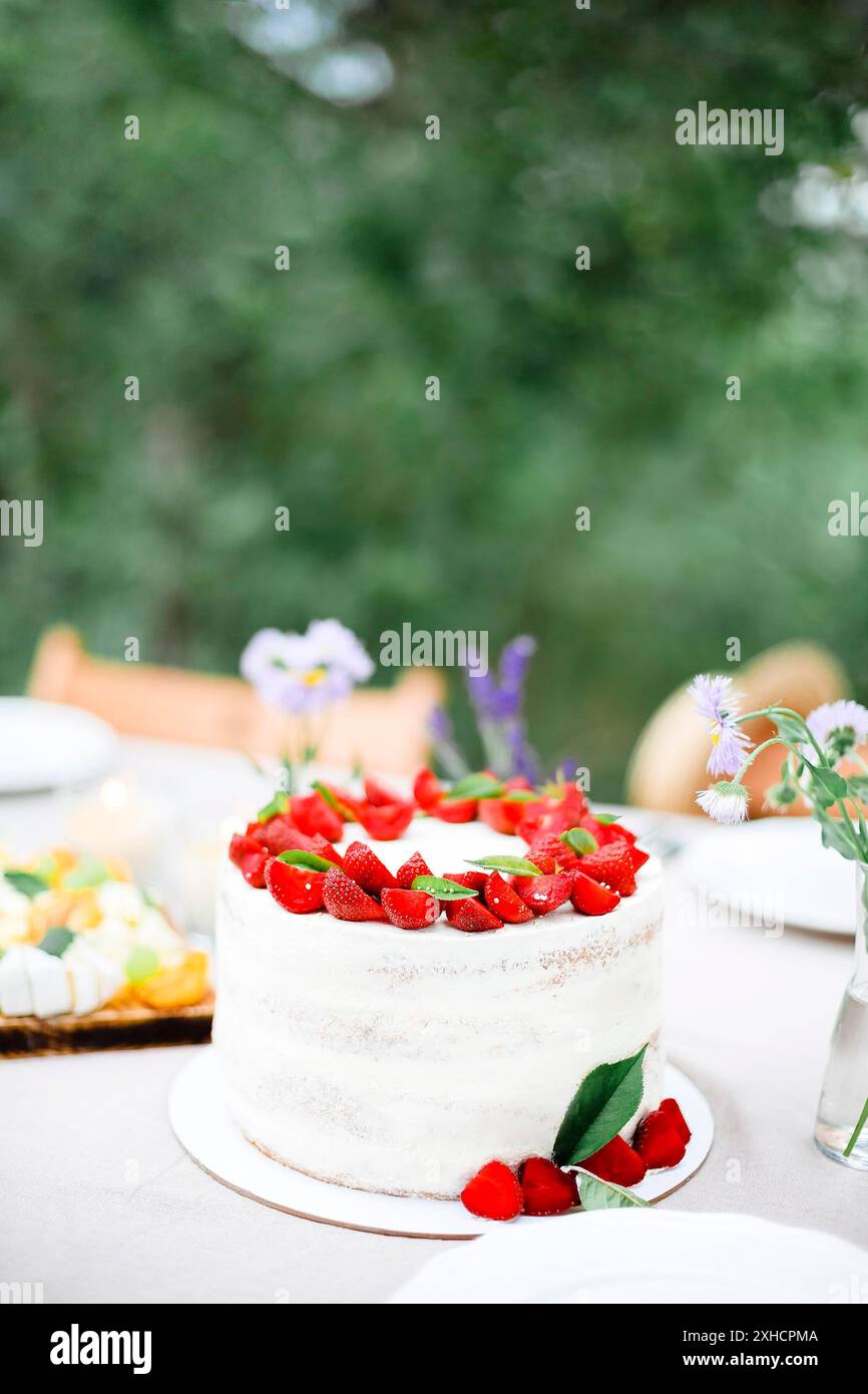 Gustosa torta con frutti di bosco posta sul tavolo da banchetto vicino a fiori e stoviglie il giorno d'estate in giardino Foto Stock