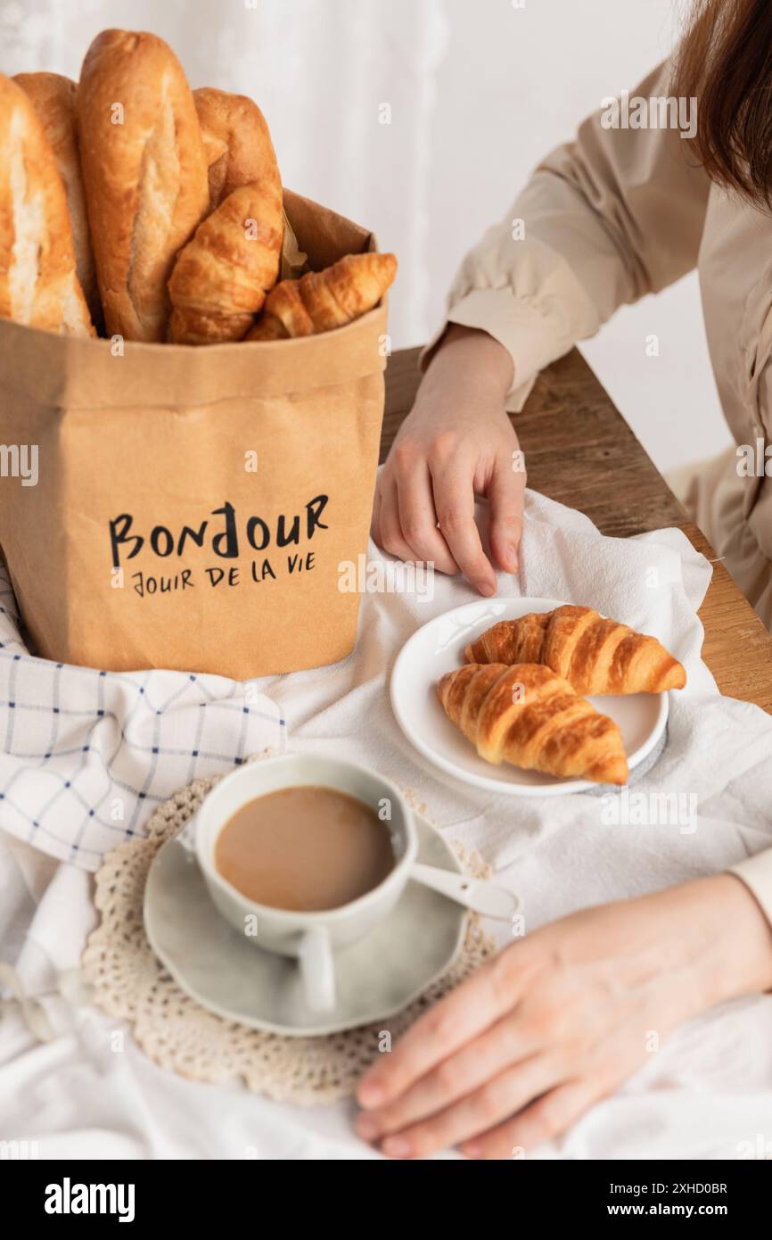 Momento delizioso: Una donna che si gusta una ciambella con caffè e pane su una tovaglia rustica. Un ananas in un sacchetto di carta kraft impermeabile. Foto Stock