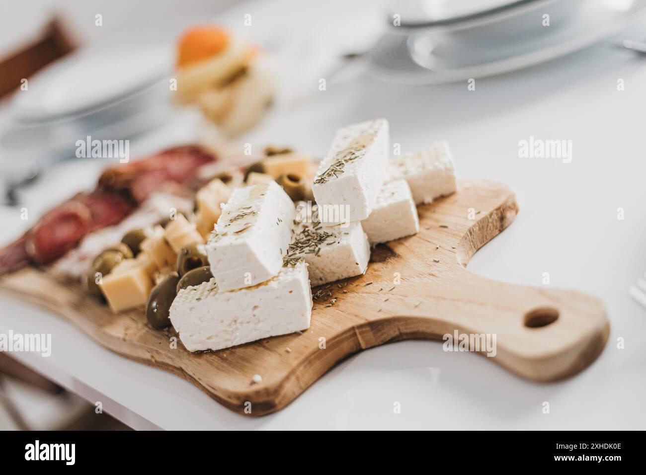 Un tagliere di legno siede su un tavolo bianco, riempito con una selezione di formaggi, olive e salumi. Foto Stock