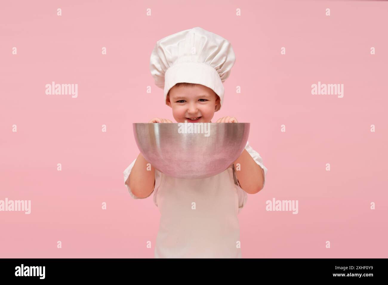 Il bambino felice, vestito da chef con cappello e grembiule, tiene una grande ciotola per un ritratto in un ambiente da studio rosa. Bambino di 3 anni (bambino di 3 anni) Foto Stock