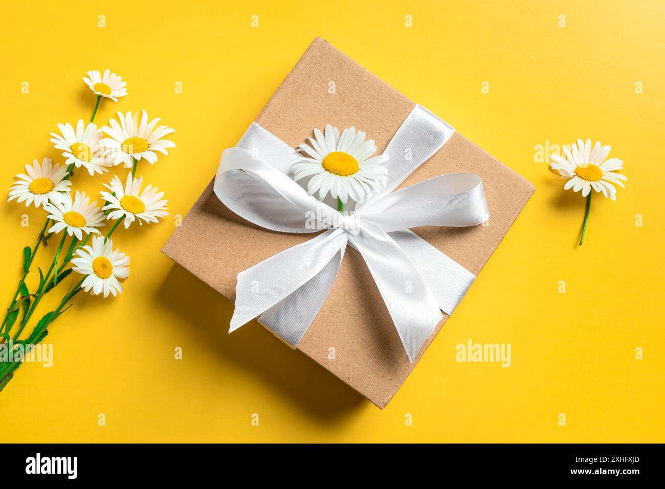 Confezione regalo con nastro bianco e mazzo di fiori camomilla su sfondo giallo brillante, vista dall'alto. Festa della mamma, compleanno e anniversario Foto Stock