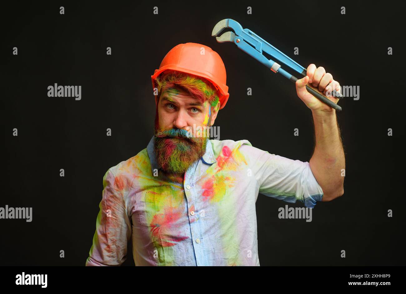 Uomo con barba in elmetto protettivo con chiave per tubi o chiave per tubazioni. Servizio di riparazione. Un idraulico o un riparatore esperto in un casco di sicurezza con chiave a piombo Foto Stock