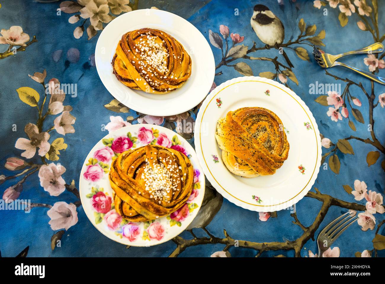 Tre piatti di classiche paste lievitate svedesi sono disposti su una tovaglia blu a motivi decorata con fiori e un uccello: kanellbullar (ciambelle alla cannella) ricoperte di zucchero perlato e kardemummaknutar (nodi al cardamomo) spolverati di semi di papavero e zucchero perlato. Entrambe le paste rientrano tra i dolci più popolari della fika svedese, la pausa caffè prolungata. Due forchette d'argento sono pronte accanto a loro. Stortorget, Stoccolma, Contea di Stoccolma, Svezia. Foto Stock