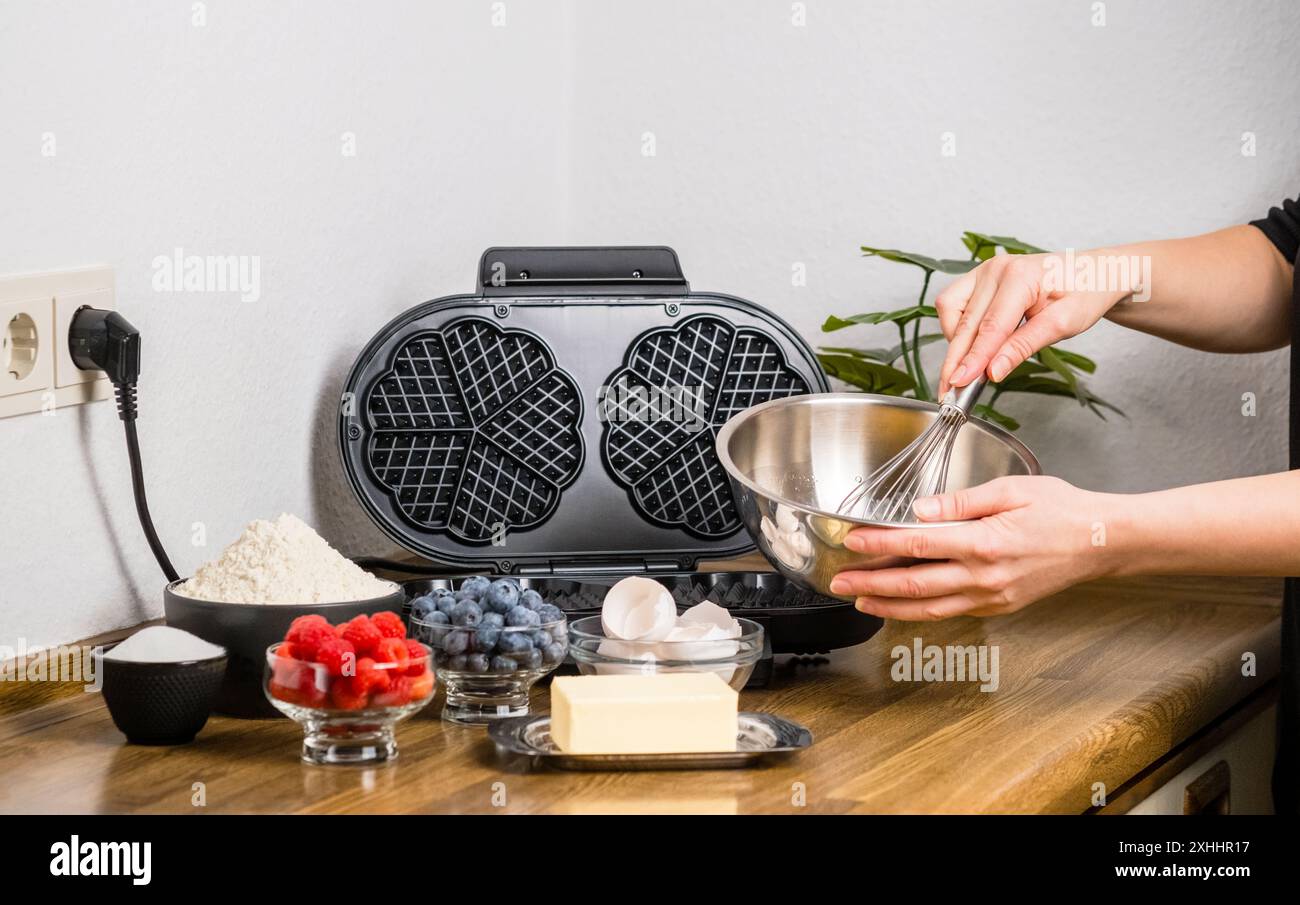 Preparare waffle belgi. Le mani delle donne sbattono le uova con una frusta in un recipiente di metallo per preparare l'impasto nella cucina di casa. Ingredienti della pastella: Farina, burro e. Foto Stock