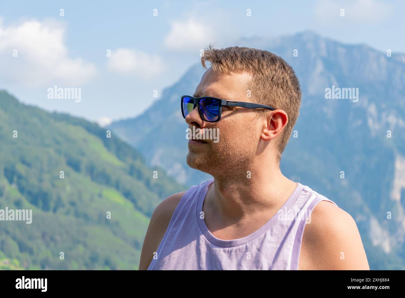 Un uomo di 40-45 anni che indossa occhiali da sole. Si trova di fronte a un lago con montagne sullo sfondo. Foto Stock