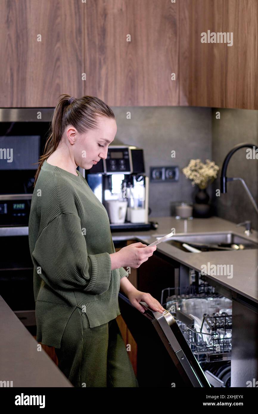 La donna ha in mano un telefono con il programma Smart Home in esecuzione, controllando a distanza la lavastoviglie. Il concetto di una cucina intelligente a casa. Foto Stock