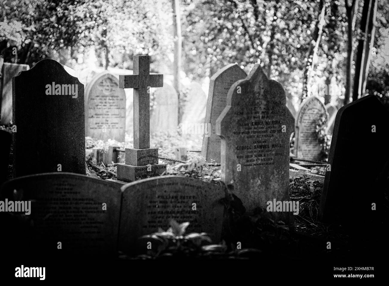 Immagine in bianco e nero di vecchie pietre tombali e croce. La croce è illuminata in un raggio di luce solare. Highgate Cemetery, Londra, Regno Unito Foto Stock