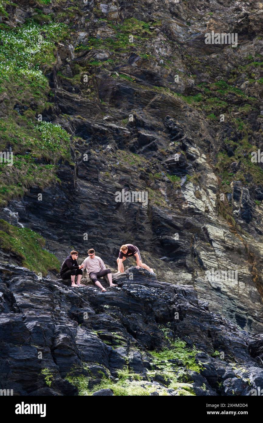 Tre vacanzieri maschi che si arrampicano su rocce sotto scogliere instabili sulla costa di Newquay in Cornovaglia nel Regno Unito. Foto Stock