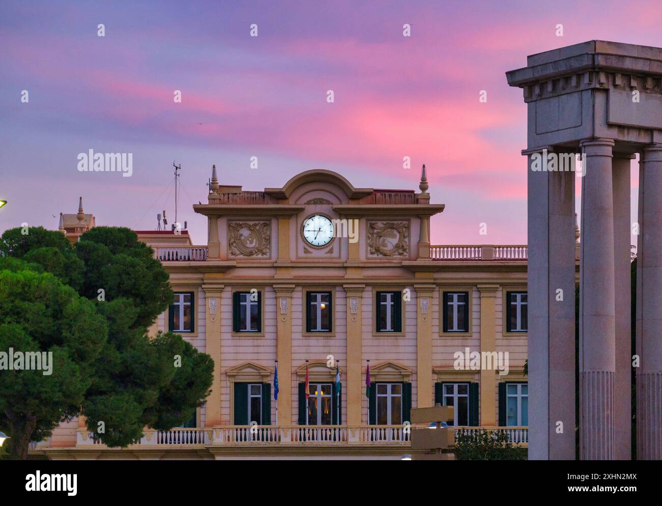 Edificio spagnolo in stile europeo con orologio, immerso nella calda luce del tramonto dell'alba, tra alberi verdi e grandiosa architettura. Foto Stock