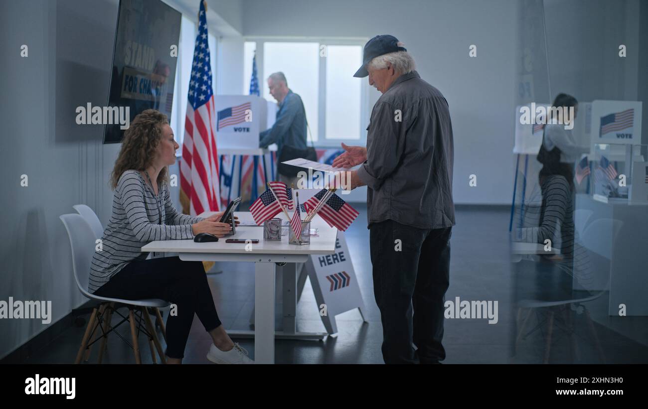 Un cittadino americano maturo del Caucaso si trova vicino al tavolo di registrazione al polling station. Una donna che ha votato parla con un uomo, usa un computer tablet e dà le schede elettorali per votare. Il giorno delle elezioni negli Stati Uniti. Foto Stock