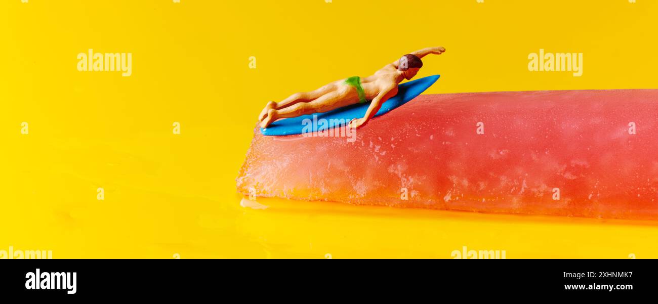 un surfista in miniatura, che indossa un costume da bagno verde, è disteso su una tavola da surf posta su uno sciogliente pop, su uno sfondo giallo, in forma panoramica Foto Stock