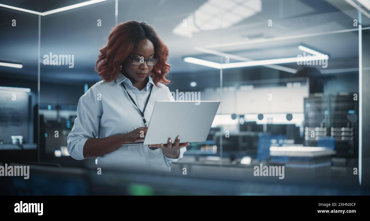 Ritratto di un responsabile del dipartimento di ingegneria del software tecnologico che sta in piedi e utilizza un computer portatile. Donna africana che guarda in lontananza e pensa mentre lavora in ufficio Foto Stock