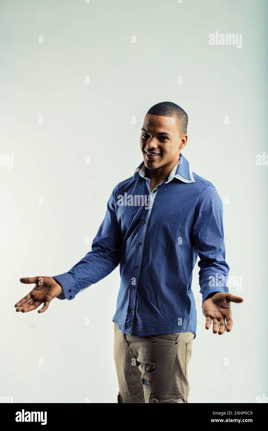 Giovane con camicia a righe blu e pantaloni beige, in piedi con le braccia aperte e le palme sollevate. La sua espressione amichevole e il suo abbigliamento informale suggeriscono che è invit Foto Stock
