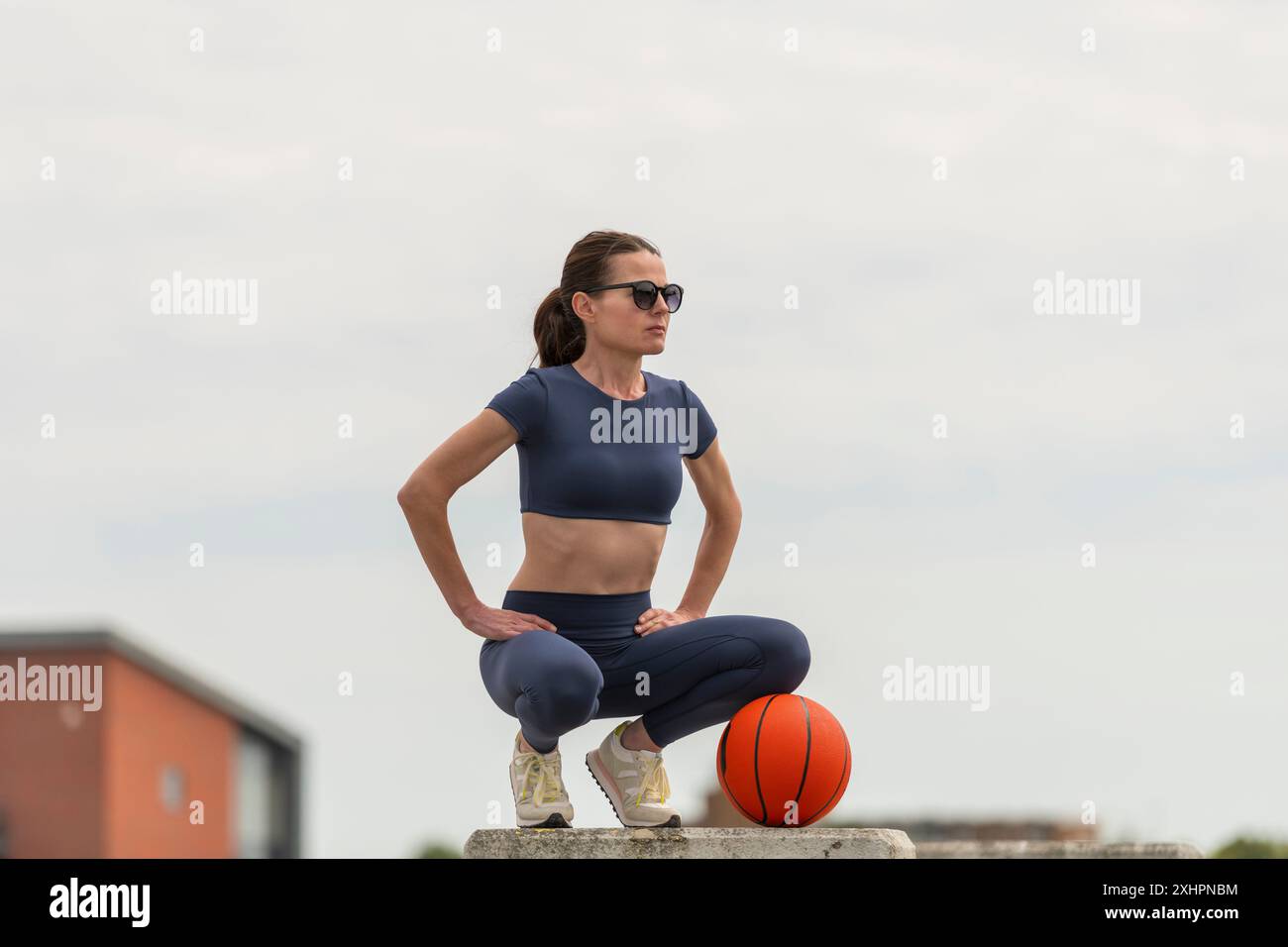 Indossate una donna sportiva che si inginocchia con un basket, un allenamento all'aperto, un ambiente urbano Foto Stock