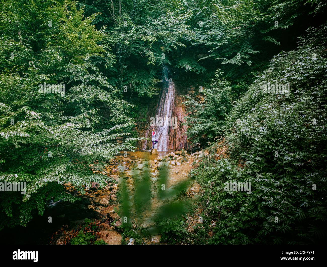 [Alto come il cielo] Una ragazza sola tra gli alberi verdi e di fronte alla cascata riceve energia positiva dalla natura facendo yoga. Foto Stock