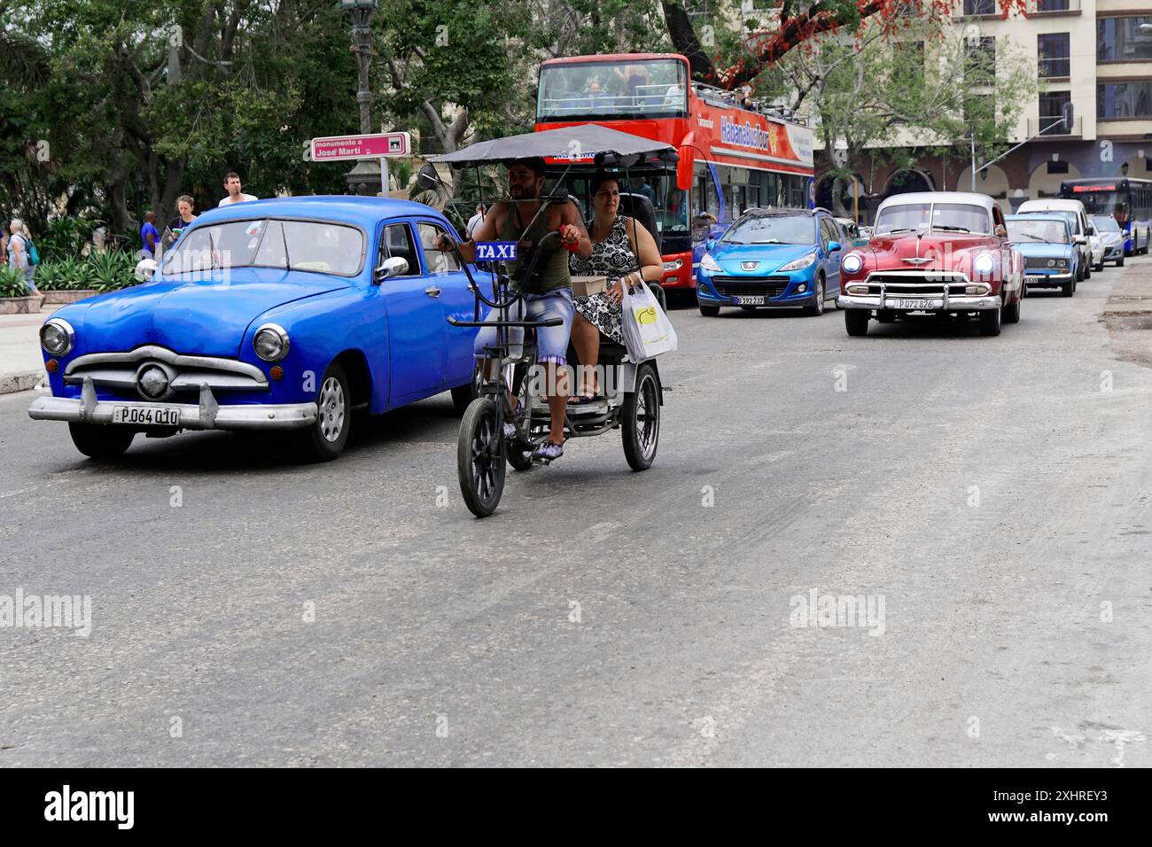 L'Avana, Cuba, America centrale, auto d'epoca blu e veicoli moderni su una strada, persone su un taxi in bicicletta Foto Stock