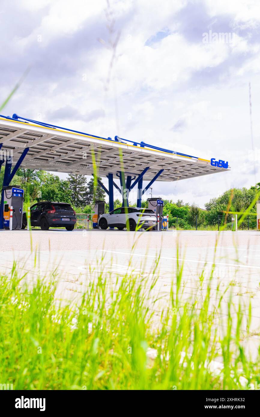 Primo piano dell'erba, dietro di essa stazioni di ricarica EnBW con tetto ad energia solare e auto elettriche, parco di ricarica ENBW, Rutesheim, Germania Foto Stock