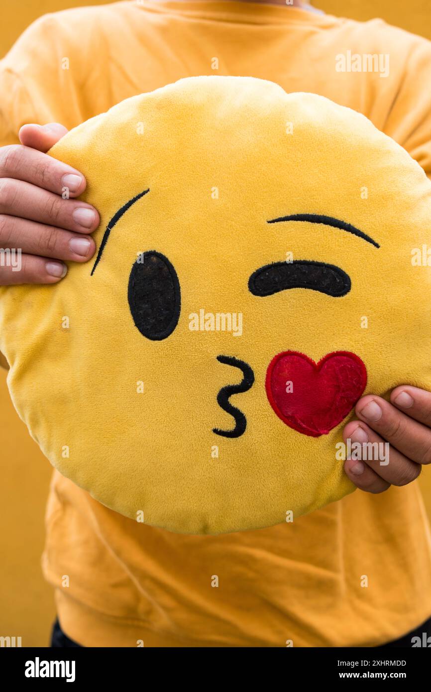Uno scatto verticale ravvicinato di una persona che tiene in mano un cuscino giallo di emoji con un viso e un cuore che bacia, indossando una camicia gialla abbinata Foto Stock