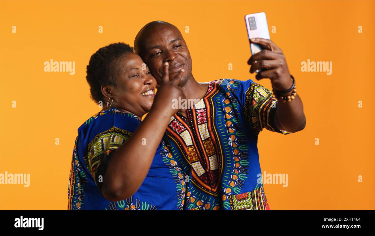 Coppia etnica sorridente che scatta foto con il telefono in studio, divertendosi a fare ricordi con la fotocamera dello smartphone. Le persone allegre scattano foto mentre indossano abiti tradizionali. Foto Stock