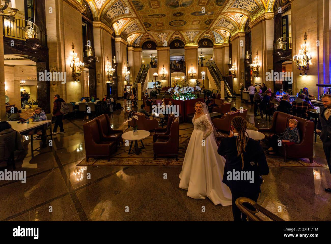Una sposa posa nel lussuosa lobby del Palmer House Hilton Hotel a Chicago, Illinois, Stati Uniti, mentre un accompagnatore sistema il suo strascico. Volte a cassettoni dorati con affreschi affrescati si estendono sopra la sala storica dell'hotel, il cui Grand Salon è stato uno dei monumenti più famosi della città dalla sua apertura nel 1873. Foto Stock
