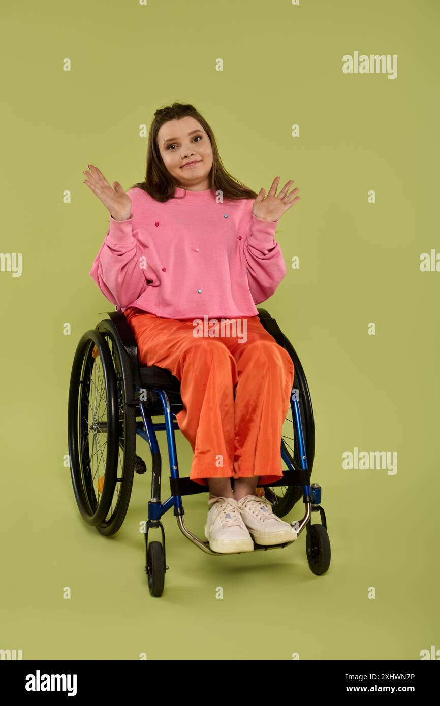 Una giovane donna con i capelli marroni siede su una sedia a rotelle in un ambiente studio. Indossa una felpa rosa e pantaloni arancioni. Foto Stock